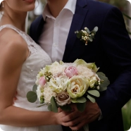 Narzeczeni obejmują się, panna młoda w białej sukni trzyma pastelowy bukiet, pan młody w granatowym garniturze z butonierką, co budzi romantyczne skojarzenia i radość.