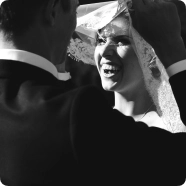 Czarno-białe zdjęcie śmiejącej się panny młodej, której twarz częściowo zakrywa koronkowy welon trzymany przez pana młodego. Scena przepełniona jest radością i światłem słonecznym.