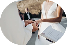 Pan młody zakłada obrączkę na palec panny młodej podczas ceremonii ślubnej na świeżym powietrzu. Urzędnik stanu cywilnego trzyma tekst ceremonii, a goście są zamazani w tle.