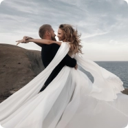 Para obejmuje się na wietrznym klifie nad morzem, a długa biała suknia kobiety powiewa dramatycznie. Scena sprawia wrażenie romantycznej i spokojnej.