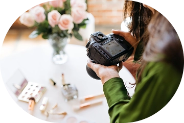 Osoba w zielonej koszuli trzyma aparat fotograficzny, kierując go w stronę stołu z kosmetykami do makijażu. W tle stoi wazon z różowymi różami. Jasna i kreatywna atmosfera.