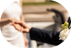 Pan młody w ciemnym garniturze z białą różą w butonierce trzyma się za ręce z panną młodą w białej sukni. Scena oddaje romantyczną, radosną chwilę.